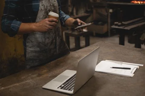 Blacksmith having coffee while using mobile phone in workshop Stock Photos