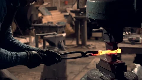 Blacksmith holding hot iron bar working with a large industrial hammer Stock Footage 132653070
