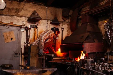 The blacksmith manually forging the molten metal on the anvil in smithy. Stock Photos