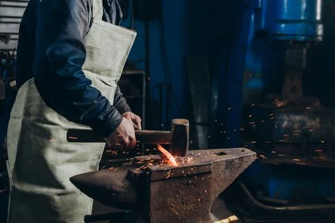 The blacksmith manually forging the red-hot metal on the anvil in smithy. Stock Photos