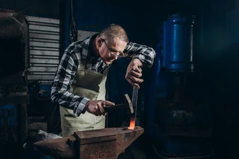 The blacksmith manually forging the red-hot metal on the anvil in smithy. Stock Photos