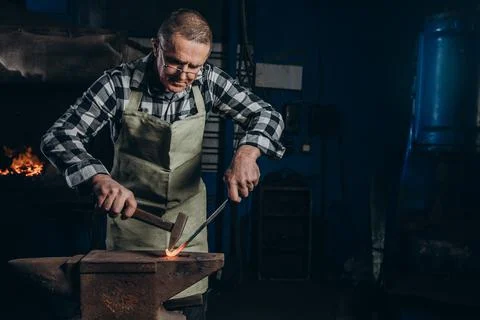 The blacksmith manually forging the red-hot metal on the anvil in smithy. Stock Photos