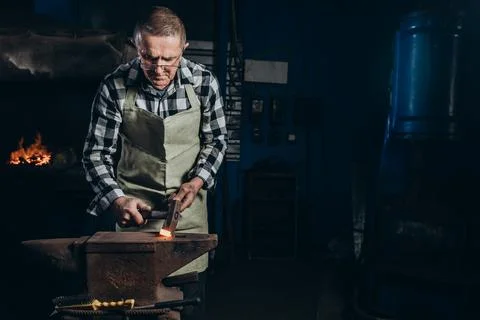 The blacksmith manually forging the red-hot metal on the anvil in smithy. Stock Photos