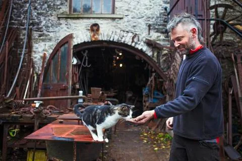 Blacksmith standing outside his workshop feeding a cat Stock Photos