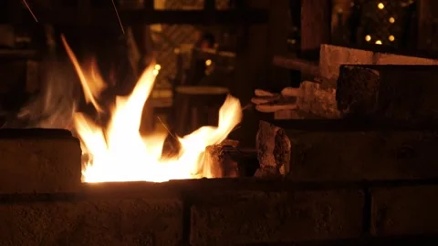 Blacksmith Tending Forge Fire with Rising Sparks in Dimly Lit Workshop Setting Stock Footage 337694490