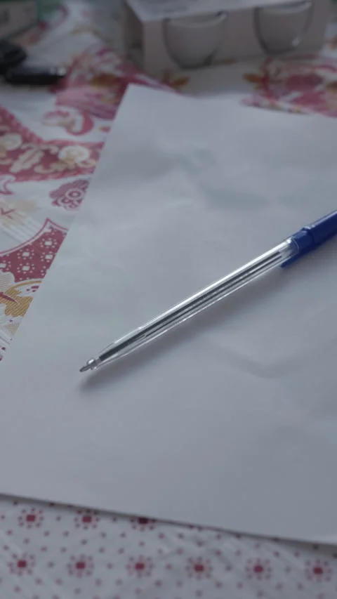 Blank paper sheet with blue ballpoint pen resting on patterned tablecloth in Stock Footage 344824673