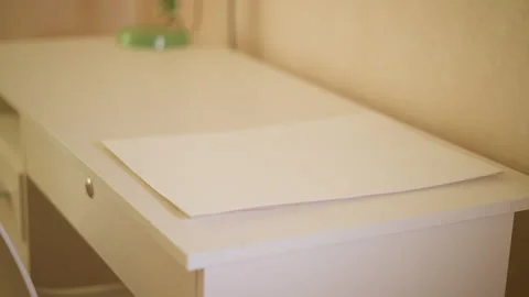 Blank white sheet of paper resting on softly lit desk with warm indoor setting Stock Footage 347229415