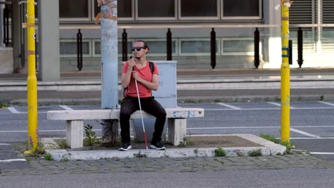 Blind man sitting on a bench undecided whether to cross the road Stock Footage 156896619
