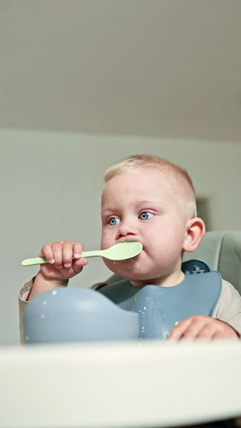 A blond boy of 2 years sits in a high chair with a silicone apron and eats Stock Footage 315537563