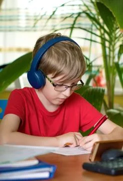 Blond boy does homework at the table in headphones Stock Photos