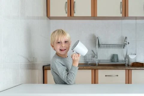 Blond boy drinking from large cup in kitchen. Funny schoolboy breakfast Фото