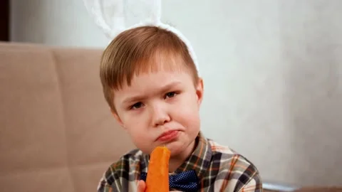 A blond boy with Easter bunny ears nibbles a carrot and pretends to be a rabbit Stock Footage 170059630