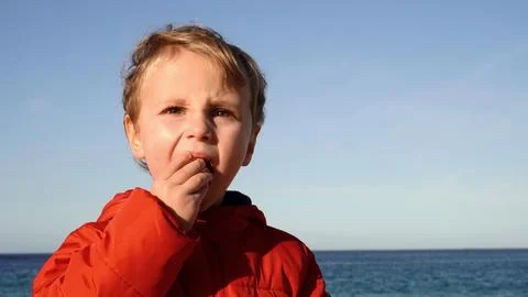 Blond boy eats a tangerine on the background of the sea. Stock Photos