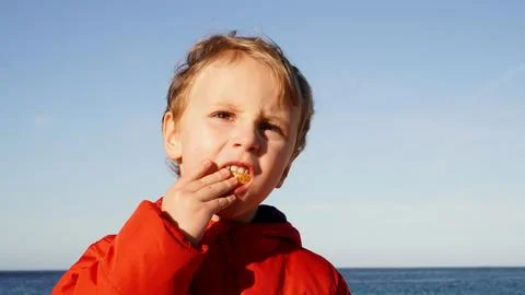 Blond boy eats a tangerine on the background of the sea. Stock Photos