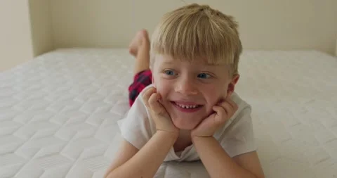 A blond boy lying on a bed in red pajama pants and a white T-shirt looks off to 스톡 동영상 324824973