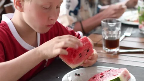 Blond boy in a red T-shirt eats a piece of watermelon at a table in a summer Stock Footage 285091634