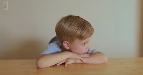 A blond boy is resting his head on his hands on the table, wearing a blue T 스톡 동영상 320646861