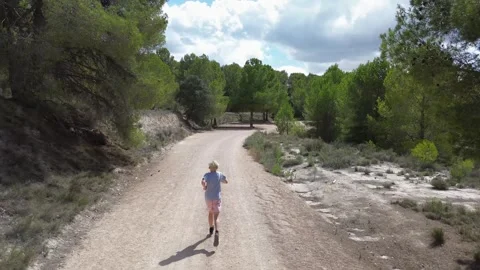 A blond boy runs along a mountain forest road, shooting on a drone from the back Stock Footage 286702374