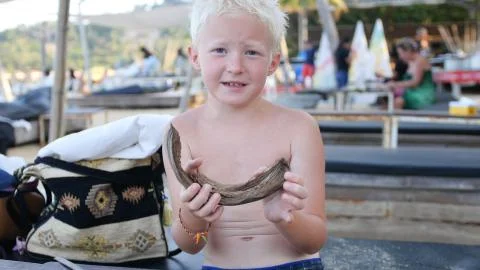 A blond boy is sitting in a cafe on the beach, playing with a stick and peeling 스톡 사진