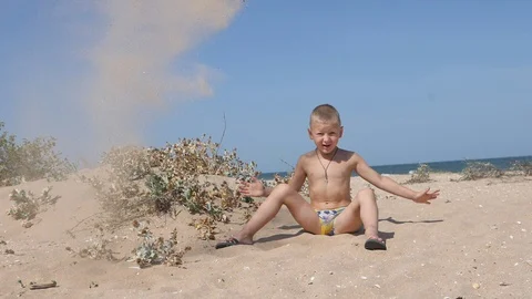 Blond boy sitting on the sandy beach throws up over his head the yellow sand. Stock Footage 99463480