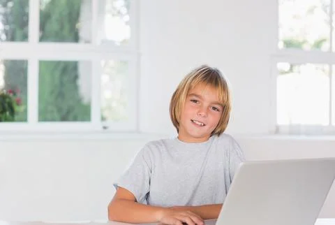 Blond child boy sitting at table using silver laptop in bright home study with Stock Photos