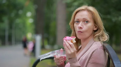Blond girl in a park eating a donut. Woman outdoors chews sweet bun Stock Footage
