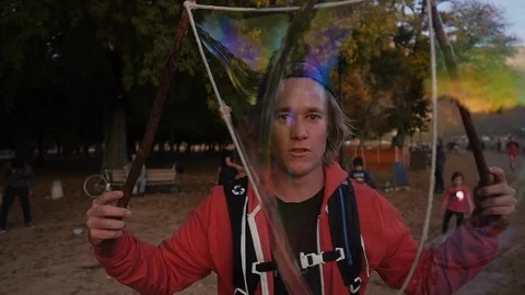Blond guy with sweatshirt making big bubble in a park with trees and people Stock Footage 87520235