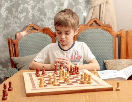 A blond-haired boy in front of a chess board with pieces, pondering a task from Stock Photos