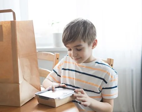 A blond-haired boy is holding a container of food, next to a delivery package on Stock Photos
