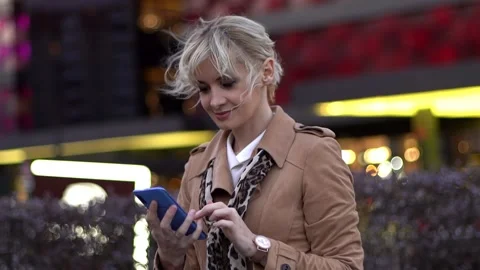 A blond-haired female using mobile phone in the urban street in early evening Stock Footage 153459490