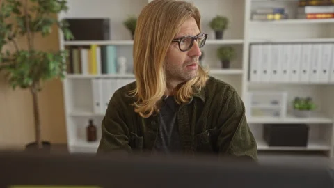Blond man rests hand on cheek at office desk with computer monitor visible;.. Stock Footage 314643356