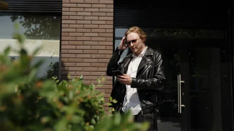 A blond man stands outside the building and looks at his phone. Sunglasses on a Stock Footage 247291474
