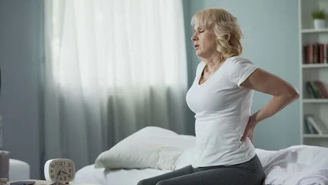 Blond mature lady sitting on bed and touching her back, radiculitis and pain Stock Footage