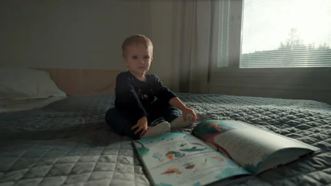 Blond toddler sits on soft bed near open book in bedroom Stock Footage 228301035
