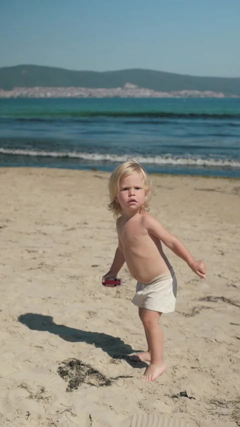Blond toddler walking toward camera on sunlit sandy beach Stock Footage 339460183