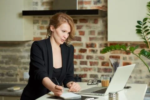 A blond woman doing notes in the notebook during a video conference at home. Stock Photos
