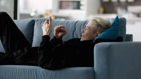 Blond young man is relaxing on grey sofa, using his gadget, smartphone, texting Stock Photos