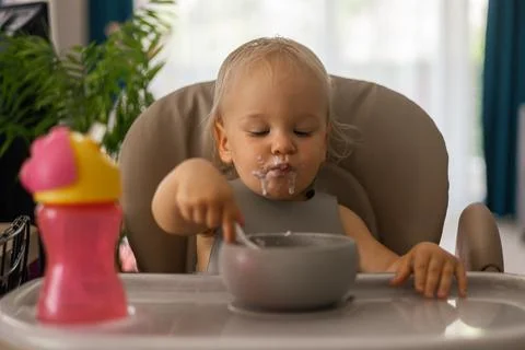 A blonde baby eats with a spoon while sitting at a children's dining table Foto stock