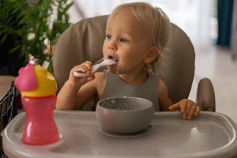 A blonde baby eats with a spoon while sitting at a children's dining table Stock Photos