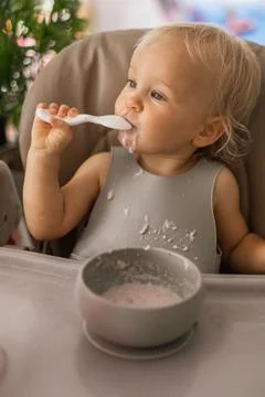 A blonde baby eats with a spoon while sitting at a children's dining table Foto stock