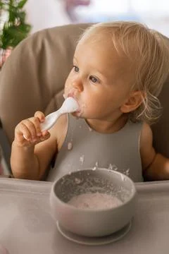 A blonde baby eats with a spoon while sitting at a children's dining table Stock Photos