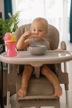 A blonde baby eats with a spoon while sitting at a children's dining table Stock Photos
