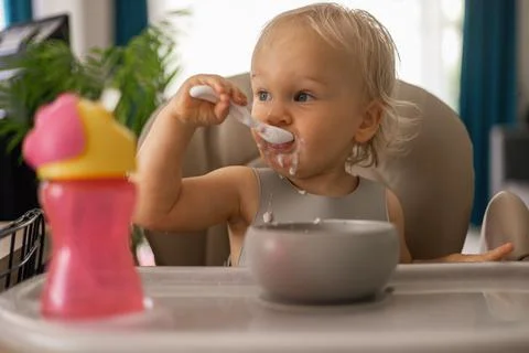 A blonde baby eats with a spoon while sitting at a children's dining table Stock Photos