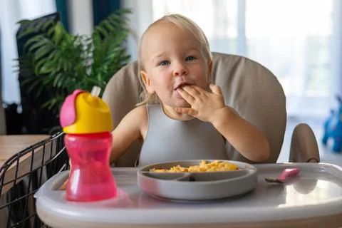 A blonde baby eats with a spoon while sitting at a children's dining table Foto stock