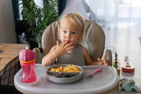 A blonde baby eats with a spoon while sitting at a children's dining table Foto stock