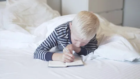 The blonde boy lies in bed with a notebook and a pen in the morning Stock Footage 123398850