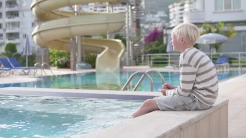A blonde boy sits on the edge of the pool with his legs lowered into the water Stock Footage 241410321