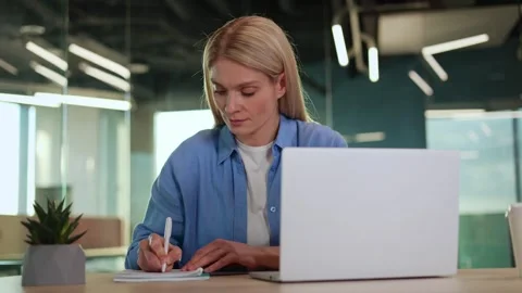 Blonde female using portable computer and notepad while sitting by table Video stock 265054812