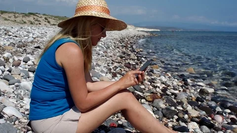 A blonde girl with hat using her mobile phone at beach Stock Footage 108232613