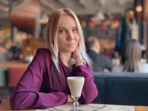 A blonde girl in a lilac sweater sits in a cozy restaurant on a sofa and she Stock Photos